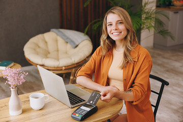 Young fun woman hold bank payment terminal to process acquire credit card pay bill sitting alone at...