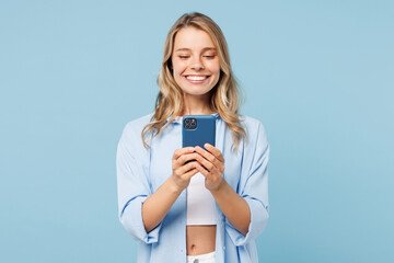Young smiling happy woman she wear white top shirt casual clothes hold in hand use mobile cell phone chat online isolated on plain pastel light blue cyan background studio portrait. Lifestyle concept