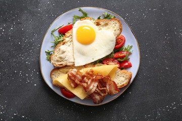 Plate with tasty fried egg, cut croissant, bacon, tomatoes and arugula on dark background