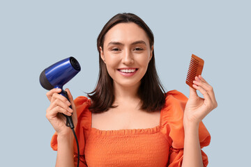 Beautiful young woman with hair dryer and comb on light background