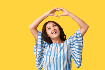 Beautiful young Asian woman showing heart gesture on yellow background. Valentine's Day celebration