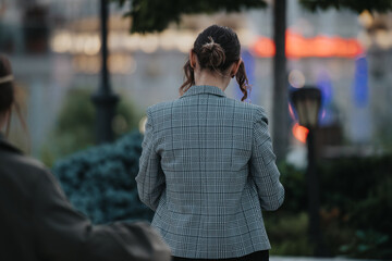 Businesswoman in a checkered blazer seen from behind, walking outdoors in a city environment with a blurred background.