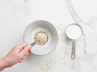 Top view of a person's hand with a measuring cup full of oat flakes over a bowl preparing porridge with milk on a white table.