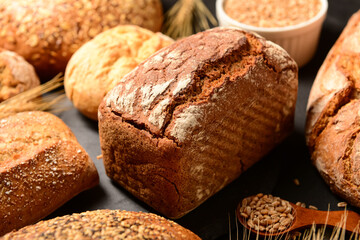 Loaves of fresh bread with buns, wheat spikelets and grains on black background
