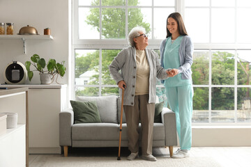 Young caregiver helping senior woman with stick to walk in kitchen