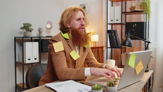 Tired exhausted frustrated bearded businessman sits in home office table with laptop sticky notes with many tasks inscription help, feels sad boredom overworked multitasking. Redhead guy freelancer.