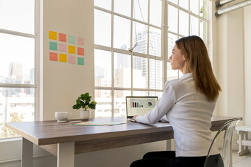 Young woman working at a desk on a computer spreadsheet while looking at notes on a wall