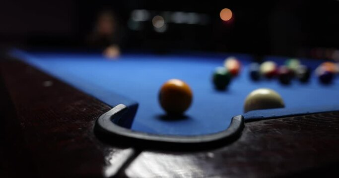 White billiard ball strikes yellow on blue billiard table. Person hits billiard ball using cue and misses hole in club dark playing room