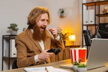 Excited Caucasian businessman eating popcorn while playing video game on laptop, spending leisure time at home office desk. Happy bearded freelancer guy playing video games with joystick controller