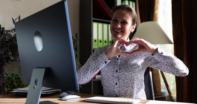Smiling woman makes heart gesture sitting at desk with computer. Happy manager shows affection and concern for colleagues and customers slow motion