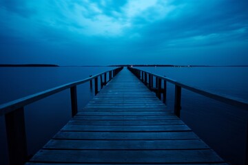 Obraz premium A tranquil image of a wooden pier stretching out into calm, deep blue waters under a cloudy twilight sky, evoking a sense of peace and solitude.