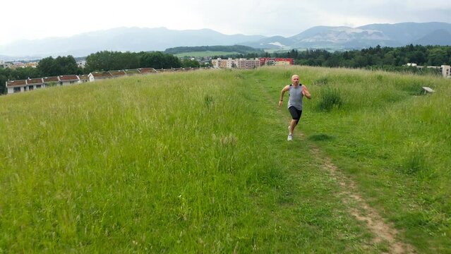 Fast running bald man on grassy trail in lush green field captured from flying drone perspective. Runner is dressed in athletic gear, showcasing active lifestyle in serene natural setting 4K video