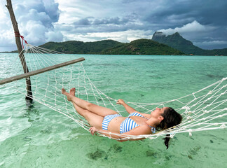 Beautiful woman relaxing in bikini swimsuit lying in rope hammock over crystal clear blue green water of Bora Bora lagoon beside Mount Otemanu while vacationing in French Polynesia