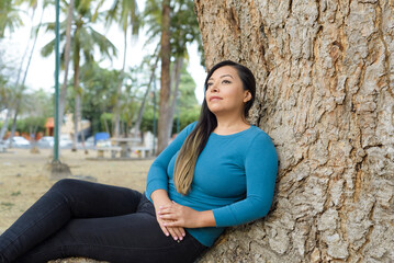 Woman seated leaning against a tree with pensive attitude.