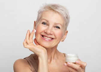 A charming elderly woman with a short hairstyle smiles and holds a jar of cream in her hands. Beauty routine.