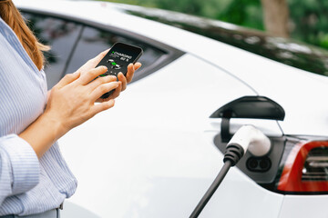 Young woman using smartphone online banking application to pay for electric car battery charging...
