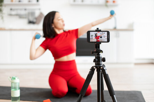 Woman in red sportswear recording fitness video at home on a smartphone. Fitness influencer demonstrating exercises with dumbbells for online audience. Healthy lifestyle and workout concept.