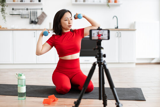 Woman in red athletic outfit exercising with dumbbells in home kitchen. Filming workout session with smartphone on tripod for online audience. Healthy lifestyle, fitness, and home workout concept.