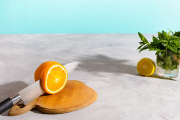 A halved orange rests on a wooden cutting board with a knife, ready for slicing, against a light blue background