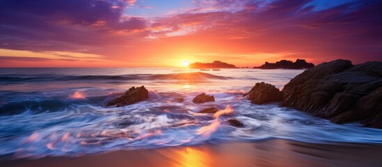 An inspirational coastal landscape features a serene beach at sunset or sunrise, with sun rays casting a colorful sky over the ocean, including a natural island and wave with white foam.