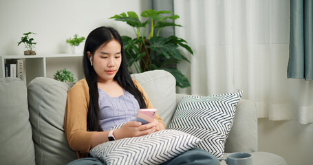 Portrait of Relaxed young Asian woman wear wireless headphones listen to music while sitting playing mobile phone on the sofa in the living room at morning ,Free time