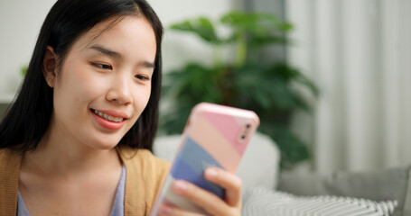 Portrait of Relaxed young Asian woman sitting on the sofa playing social media on mobile phone in the living room at morning ,Free time