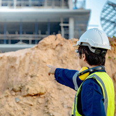 Asian soil engineer in reflective green vest with helmet, earmuffs and goggles pointing at pile of laterite soil in front of building site. Geotechnical engineering for construction base