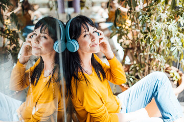 Smiling mature woman wearing wireless headphones listening to music by glass