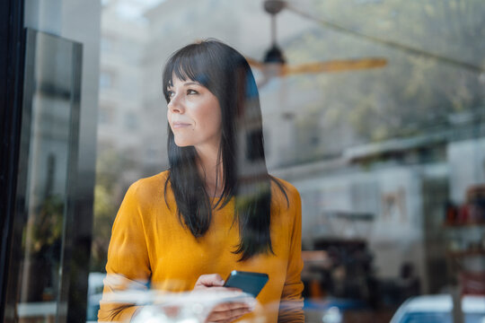 Thoughtful woman with smart phone seen through glass in cafe