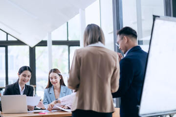 In a corporate meeting, a businessman leads a discussion on financial strategy, pointing at charts and graphs. The team collaborates on planning and analysis, using laptops and paperwork to ensure suc