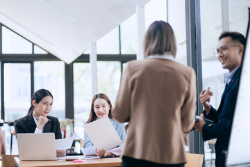 In a corporate meeting, a businessman leads a discussion on financial strategy, pointing at charts and graphs. The team collaborates on planning and analysis, using laptops and paperwork to ensure suc