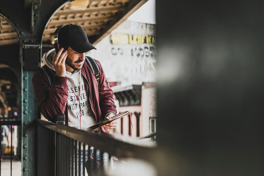 Young man holding digital tablet listening through headphones waiting at station