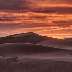 A sunset in the desert: the sand dunes are dyed a deep orange, while the sky is transformed into a canvas of orange, reddish and violet tones.