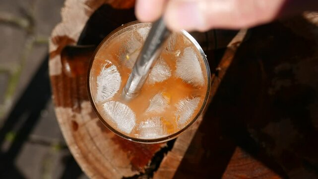 A POV of a person mixing a Caipirinha glass on the table. A Tangerine Caipirinha with cacha&ccedil;a, big ice cubes and sugar. A man stirring a Caipirinha cocktail with a metal drinking straw. A top view.