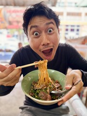 Vertical photo of asian man with wow funny face, presenting and showing Thai noodles, eating street...