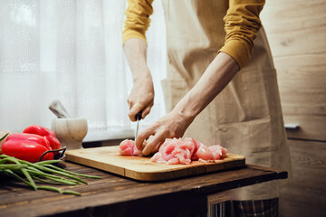 Unrecognizable man in apron slicing chicken fillet with kitchen knife in the kitchen
