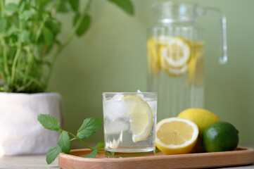 Summer lemonade drink in glass on wooden kitchen table with lemons and ice, fresh green background,...