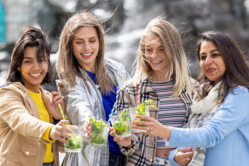 Four women enjoying nature near a waterfall, bonding outdoors, toasting with cocktails and smiling in a scenic setting