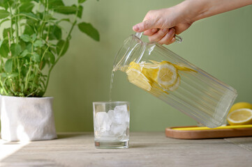 Hand holding jug with lemonade drink pouring beverage in glass with ice, soft green background, plant and lemons on kitchen table, summer non alcohol healthy drink