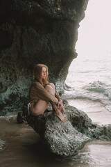 A young red-haired woman in a bikini sits on a rock against which huge ocean waves crash, sea foam rises to the top and splashes. Fashion portrait against the background of the ocean. Freedom concept.