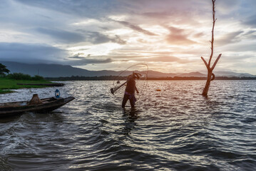 Fototapeta premium Asian fisherman on wooden boat casting a net for catching freshwater fish in nature river in the early morning before sunrise