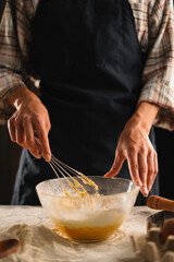 Close up of man in checkered shirt and black apron whisking flour and egg mixture in glass bowl. Man busy kneading dough creating rustic and homely atmosphere