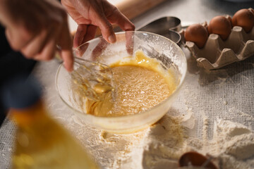 Hand whisking yellow batter in glass bowl on floured table. Nearby are brown eggs in a carton, heart-shaped cookie cutter, and rolling pin