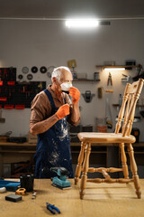 An elderly man in orange gloves, a blue apron, stands in the workshop in front of an old wooden chair, puts on a protective mask on his face. © Andrii Lysenko