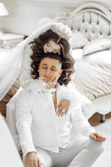 A man and woman are posing for a picture, with the woman wearing a white wedding dress and the man wearing a white shirt. The man is sitting on the bed while the woman is standing on top of him