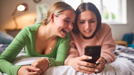 Two Teenage Girls At Home In Bedroom Laughing At Social Media Or Messages On Mobile Phone