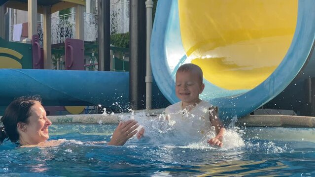 Happy child with mother have fun swimming in pool of summer resort hotel, little boy in swim vest life jacket for kids slides down water slide into pool