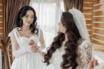 Two women are standing in a room, one of them is wearing a wedding dress. They are smiling and seem...