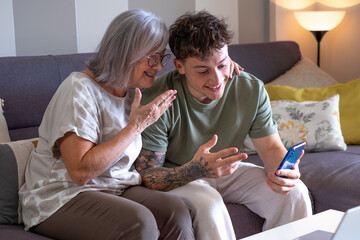 Video call concept. Smiling handsome boy and his grandmother sitting together on sofa using...