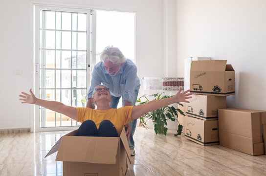 Cheerful senior couple moves into a new house, surrounded by moving boxes in an empty room, two excited seniors play with an empty box